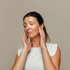 Woman applying an OmniLux HydroGel Sheet Mask to her face against a beige background