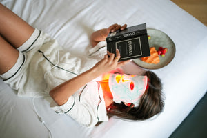 Person lying on a bed holding a Chanel book with a plate of food nearby
